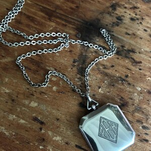 May include: Silver-toned necklace with a rectangular pendant. The pendant features a diamond-shaped design with engraved initials. The chain is made of small, interconnected links. The necklace rests on a wooden surface.