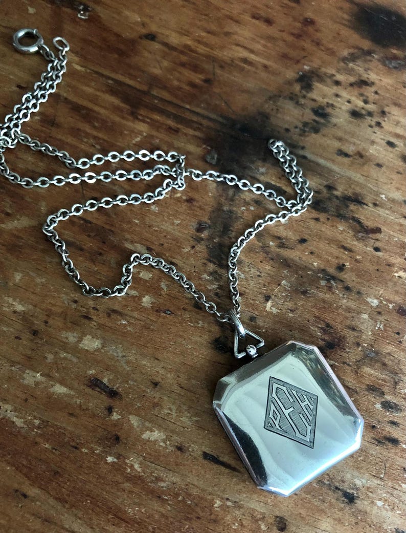 May include: Silver-toned necklace with a rectangular pendant. The pendant features a diamond-shaped design with engraved initials. The chain is made of small, interconnected links. The necklace rests on a wooden surface.