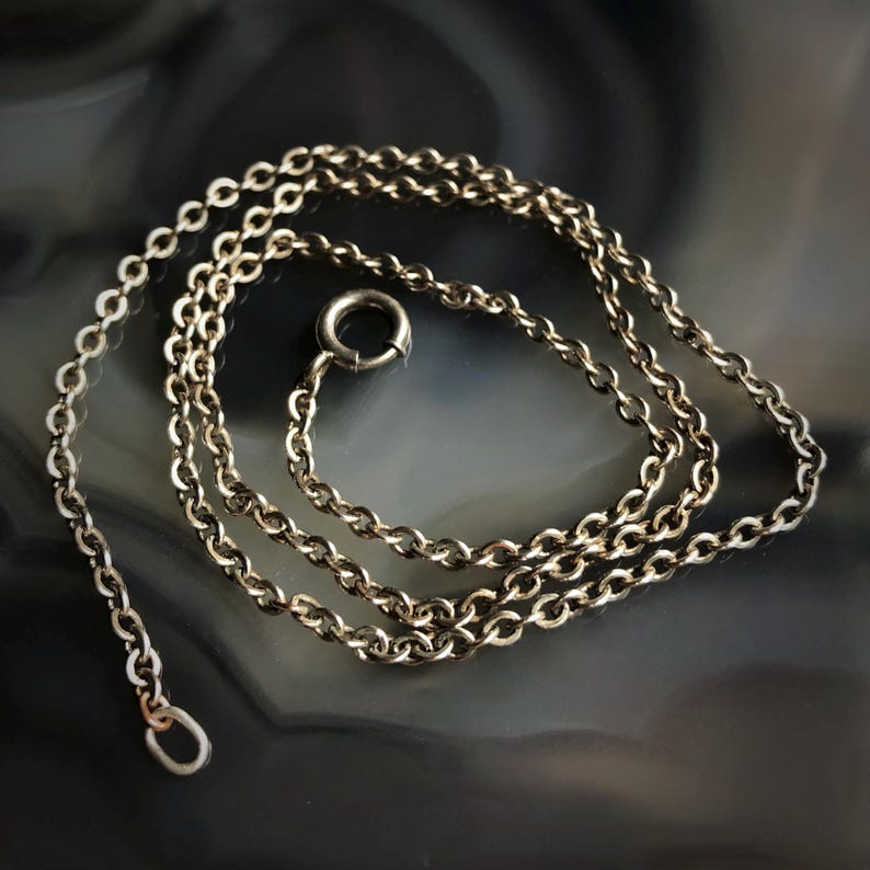 May include: A silver-colored necklace with a delicate chain design. The necklace features a small clasp and a loop at the end for attaching a pendant. The chain is made of small, interconnected links.