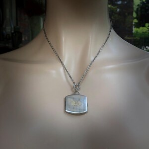 May include: Silver-toned pendant necklace with a square, slightly curved pendant. The pendant has a rectangular, light-colored center with an embossed design. The necklace chain is a delicate, linked design, and the pendant hangs from a looped bail.