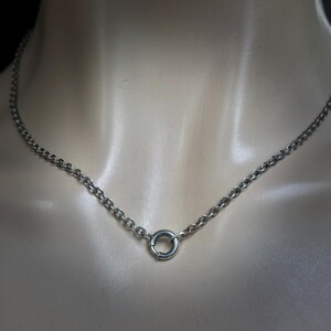 May include: A silver-colored chain necklace with a circular clasp. The necklace is composed of small, rounded links. The clasp is a small, circular ring. The necklace is displayed on a mannequin.