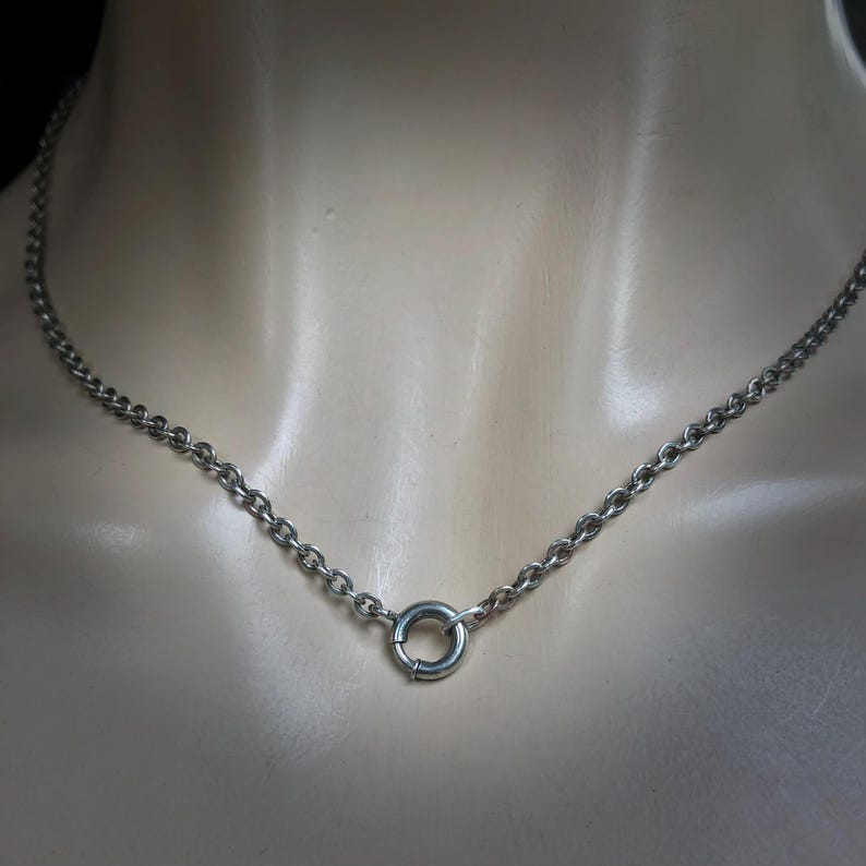 May include: A silver-colored chain necklace with a circular clasp. The necklace is composed of small, rounded links. The clasp is a small, circular ring. The necklace is displayed on a mannequin.