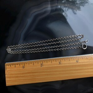 May include: A silver-colored chain necklace with a small clasp, lying on a dark, patterned surface. A wooden ruler with inch markings is positioned below the chain, showing measurements.