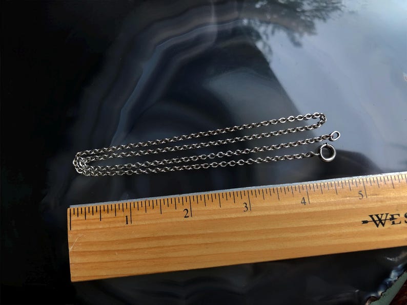 May include: A silver-colored chain necklace with a small clasp, lying on a dark, patterned surface. A wooden ruler with inch markings is positioned below the chain, showing measurements.