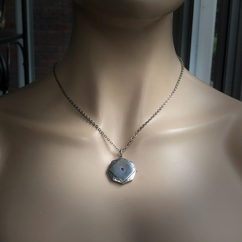 May include: Silver-toned necklace with a geometric pendant. The pendant is a square shape with a textured surface and a small, round, red gemstone in the center. The necklace is displayed on a mannequin.