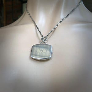 May include: A silver-toned pendant necklace. The pendant is a rectangular shape with vertical lines and an oval design with an ornate letter. The necklace chain is a linked design.