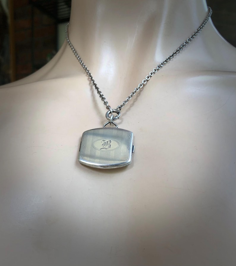 May include: A silver-toned pendant necklace. The pendant is a rectangular shape with vertical lines and an oval design with an ornate letter. The necklace chain is a linked design.