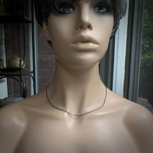 May include: A silver-colored chain necklace displayed on a mannequin. The necklace is delicate and simple, with a fine chain. The mannequin has short, dark hair and is positioned in front of a window.