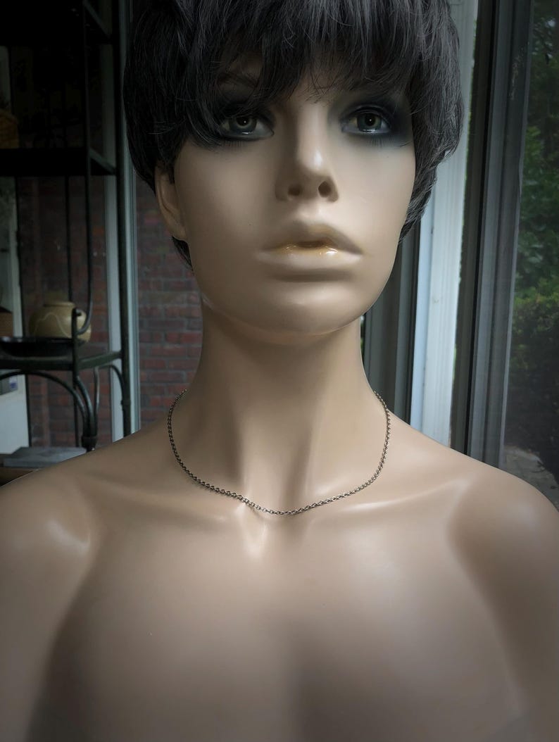 May include: A silver-colored chain necklace displayed on a mannequin. The necklace is delicate and simple, with a fine chain. The mannequin has short, dark hair and is positioned in front of a window.