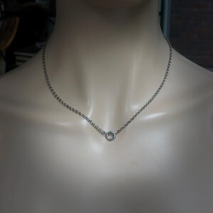May include: A silver-colored necklace with a delicate chain and a small, circular pendant. The necklace is displayed on a mannequin, showcasing its simple and elegant design. The chain is made of small, interconnected links.