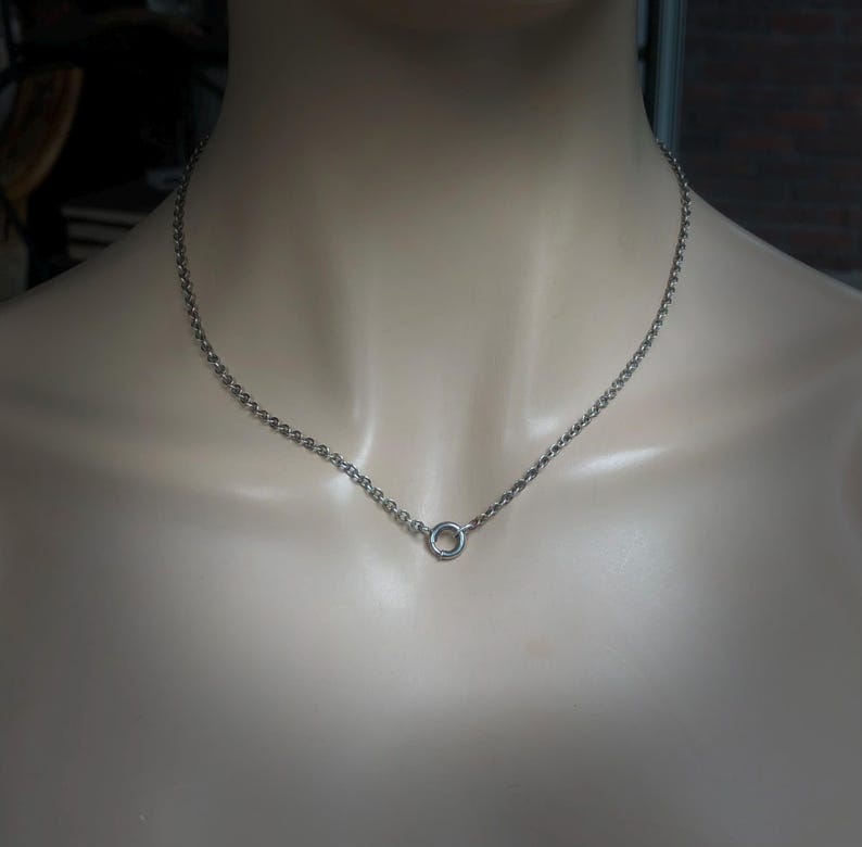 May include: A silver-colored necklace with a delicate chain and a small, circular pendant. The necklace is displayed on a mannequin, showcasing its simple and elegant design. The chain is made of small, interconnected links.