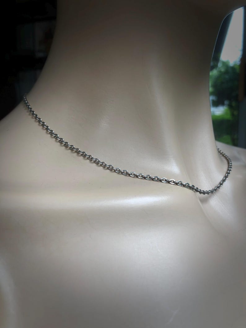 May include: A silver-colored chain necklace draped around a mannequin's neck. The chain is made of small, interconnected links, creating a delicate and simple design. The necklace is a classic accessory.