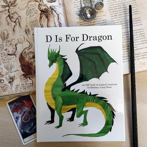 Puede incluir: Un libro infantil titulado "D Is For Dragon" con una ilustración de un dragón verde y amarillo. El libro forma parte de una serie de libros ABC sobre criaturas mágicas de Brittany Long Olsen.