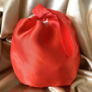 May include: A bright red satin drawstring bag, gathered at the top with a matching ribbon closure. The bag is set against a backdrop of shimmering gold satin fabric, creating a luxurious feel. The bag's design is simple yet elegant.