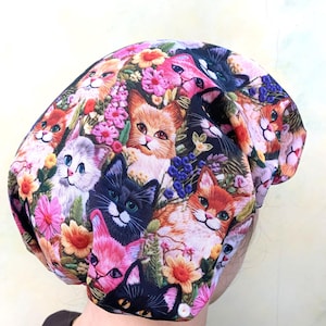 Op de afbeelding: Een kleurrijke beanie-muts met een patroon van katten en bloemen. De muts toont verschillende kattenrassen in tinten oranje, zwart, roze en wit, omringd door roze, gele en paarse bloemen. De muts is gemaakt van een zacht, flexibel materiaal.