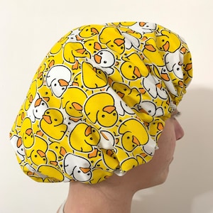 Pode incluir: Uma touca de banho amarela com um padrão repetido de patos de desenho animado amarelos, brancos e laranja. A touca é feita de tecido e foi projetada para proteger o cabelo da água durante o banho ou duche.