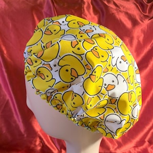 May include: A scrub cap with a repeating pattern of yellow and white cartoon ducks. The ducks have orange beaks and are set against a yellow background. The cap is designed to cover the head and hair.