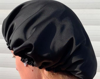 May include: A black satin bonnet, designed to protect hair. The bonnet is worn on the head, covering the hair. The material has a slight sheen. The background is a white, slatted surface.