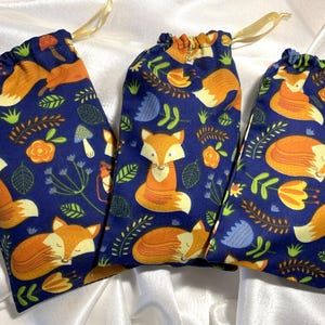 Op de afbeelding: Drie trekkoordzakken met een marineblauwe achtergrond en een herhalend patroon van oranje vossen, bloemen en bladeren. De zakken hebben een gouden trekkoordsluiting.