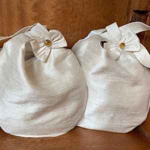 May include: Two ivory-coloured, round handbags with handles. Each bag has a bow with a decorative jewel. The bags are made of a textured fabric, and the handles are attached to the top of the bags.