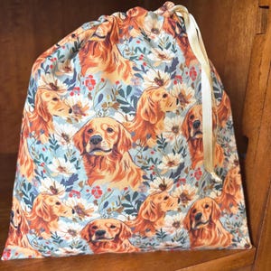 Może przedstawiać: Materiałowy worek ze ściągaczem ozdobiony powtarzającym się wzorem psów rasy golden retriever, przeplatanym białymi i pomarańczowymi kwiatami oraz zielonymi liśćmi na jasnoniebieskim tle, wykończony kremową wstążką satynową.