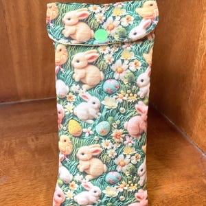 May include: A rectangular glasses case with a flap closure, decorated with an Easter-themed pattern. The fabric design includes bunnies, chicks, eggs, and flowers on a green background. The flap is secured by a green button.