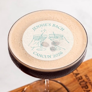 May include: A close-up of a cocktail with a white sticker on top. The sticker has a drawing of two martini glasses and the text "Jennie's Bach Cancun 2026".