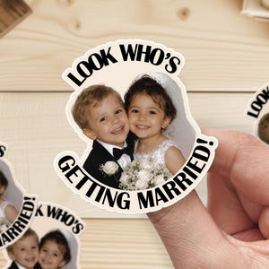 May include: A die-cut sticker featuring a photograph of a young boy in a black tuxedo and a young girl in a white wedding dress with a veil, holding a bouquet of white flowers. The text "LOOK WHO'S GETTING MARRIED!" is printed in black around the image.