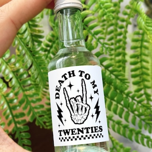 May include: A small clear glass bottle with a white label that says "Death to My Twenties". The label has a black and white illustration of a skeleton hand making a rock and roll hand gesture.