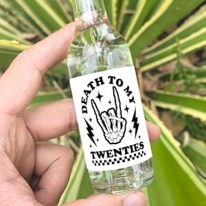 May include: A clear glass bottle with a copper-colored cap and a white label that says "Death to My Twenties" with a black and white illustration of a skeleton hand making a rock and roll hand gesture.