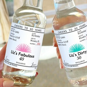 May include: Two clear glass bottles with white labels. The labels have black text that reads "Cheers to 40 Years" and "Cheers to 30 Years". The labels also have a pink and turquoise agave plant design. The text on the labels also includes "Liz's Fabulous 40" and "Liz's Dirty 30".