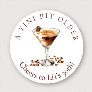 May include: A white round sticker with brown text that reads "A TINI BIT OLDER Cheers to Liz's 30th!"  A brown illustration of a martini glass with coffee beans around it.