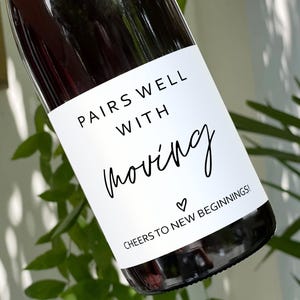 May include: A white wine bottle with a label that says "Pairs well with moving. Cheers to new beginnings!" The label has a small heart symbol.