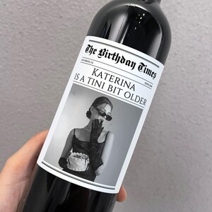 May include: A black and white photo of a woman wearing sunglasses and a black dress holding a cake. The photo is on a wine bottle label with the text "The Birthday Times" and "Katerina is a Tini Bit Older".