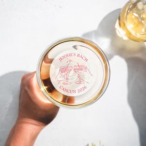 May include: A white round sticker with pink text that reads "Jennie's Bach Cancun 2026". The sticker features a line drawing of two martini glasses with stars above them.