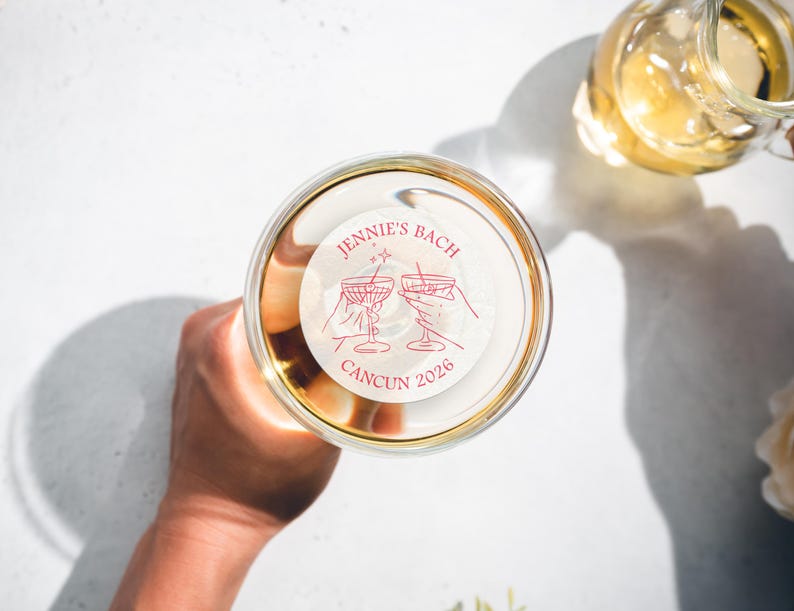 May include: A white round sticker with pink text that reads "Jennie's Bach Cancun 2026". The sticker features a line drawing of two martini glasses with stars above them.