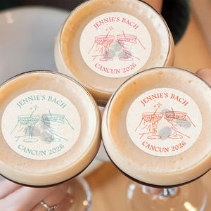 May include: Three clear glass cups with a beige liquid inside. Each cup has a round sticker with the text "JENNIE'S BACH CANCUN 2026" and a graphic of two martini glasses with stars above them.