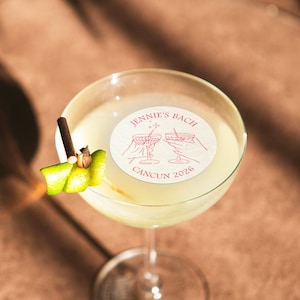 May include: A clear glass cocktail glass with a yellow drink and a lime garnish. A white sticker with the text "Jennie's Bach Cancun 2026" and a pink illustration of two martini glasses is on the side of the glass.