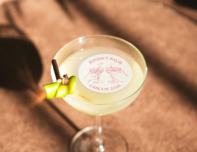 May include: A clear glass cocktail glass with a yellow drink and a lime garnish. A white sticker with the text "Jennie's Bach Cancun 2026" and a pink illustration of two martini glasses is on the side of the glass.