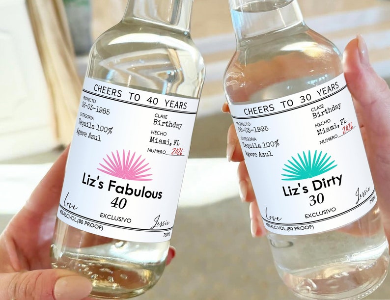 May include: Two clear glass bottles with white labels. The labels have black text that reads "Cheers to 40 Years" and "Cheers to 30 Years". The labels also have a pink and turquoise agave plant design. The text on the labels also includes "Liz's Fabulous 40" and "Liz's Dirty 30".
