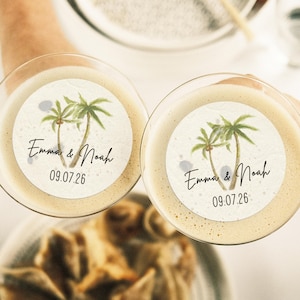 May include: Two clear glass cocktail glasses with paper drink coasters. The coasters feature a tropical palm tree design and the text "Emma & Noah 09.07.26".