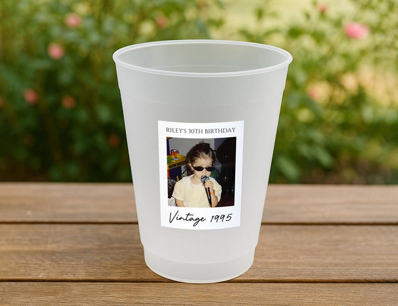 May include: A frosted plastic cup with a sticker featuring a vintage photo of a child wearing sunglasses and holding a microphone. The sticker reads "Riley's 30th Birthday" and "Vintage 1995". The cup is sitting on a wooden surface.