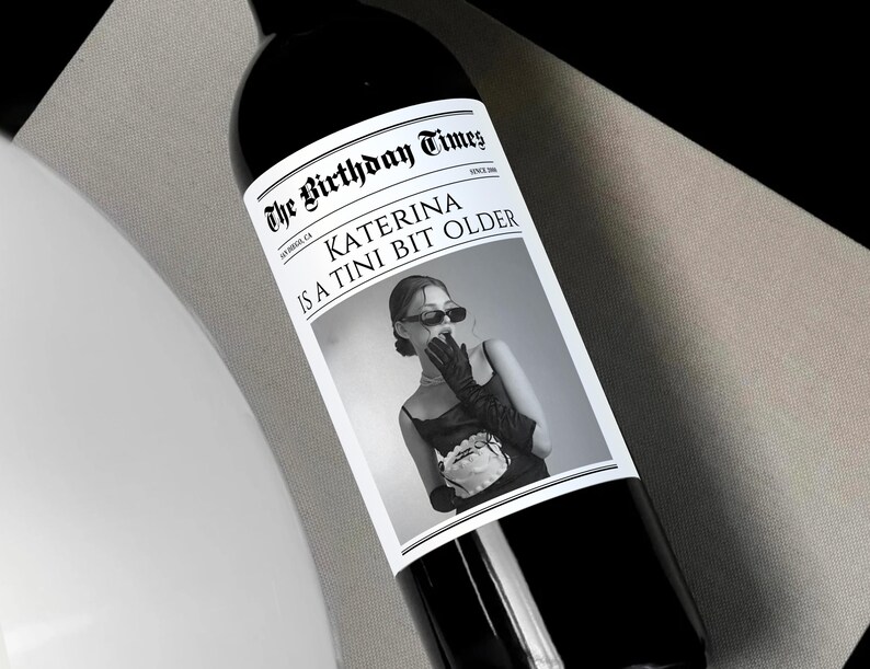 May include: A black and white photo of a woman with a surprised expression on her face. The photo is on a wine bottle label with the text "The Birthday Times" and "Katerina is a Tini Bit Older".
