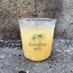 May include: A clear plastic cup filled with a yellow beverage, possibly lemonade or juice. The cup has a sticker with two palm trees, the names "Emma & Noah", and the date "09.07.26". The cup is sitting on a rough, gray stone surface.