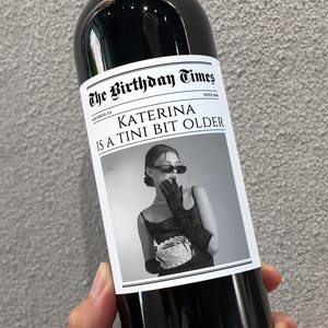 May include: A black and white photo of a woman wearing a black dress and gloves on a wine bottle label. The label reads "The Birthday Times" and "Katerina is a Tini Bit Older".