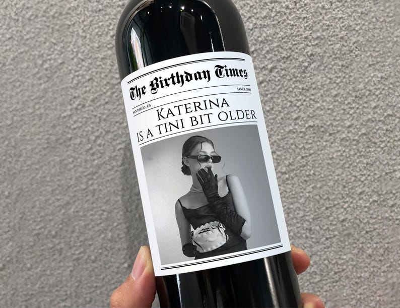 May include: A black and white photo of a woman wearing a black dress and gloves on a wine bottle label. The label reads "The Birthday Times" and "Katerina is a Tini Bit Older".