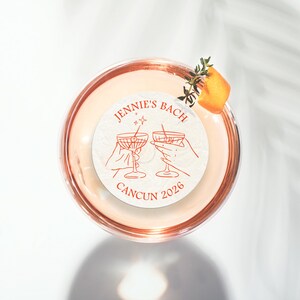 May include: A white round sticker with the text "Jennie's Bach Cancun 2026" and a graphic of two hands holding up martini glasses. The sticker is on top of a clear glass with a pink liquid inside.