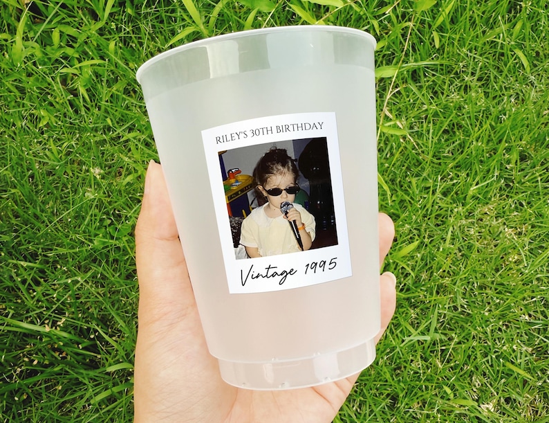 May include: A frosted plastic cup with a photo label that reads "Riley's 30th Birthday" and "Vintage 1995." The photo shows a young person wearing sunglasses and holding a microphone. The cup is held against a background of green grass.
