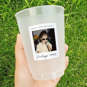 May include: A frosted plastic cup with a photo label that reads "Riley's 30th Birthday" and "Vintage 1995." The photo shows a young person wearing sunglasses and holding a microphone. The cup is held against a background of green grass.