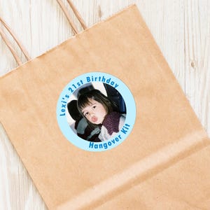Op de afbeelding: Een ronde blauwe en witte sticker met een foto van een jong persoon in een auto. De tekst op de sticker luidt "Lexi's 21st Birthday Hangover Kit".